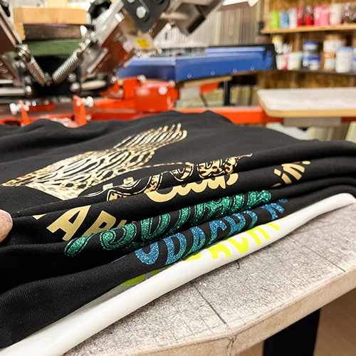 カラフルなデザインがプリントされた複数のTシャツと工房の印刷機材